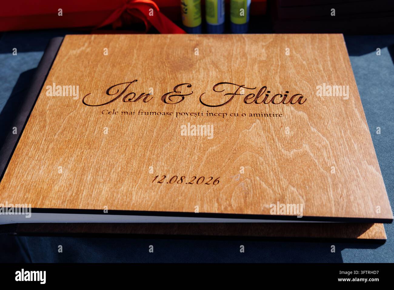 Libro degli ospiti in legno con nomi incisi Jon & Felicia data 12.08.2026 primo piano che mostra la calda venatura del legno e l'incisione dello script sulla copertina appoggiata su uno sfondo scuro Foto Stock