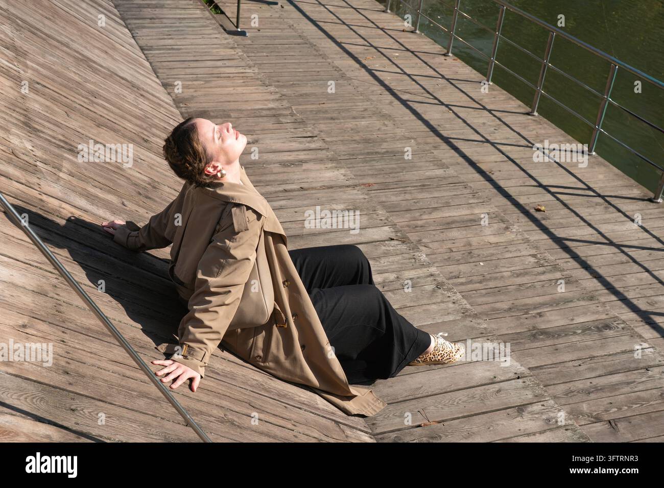 Donna si appoggia all’indietro sui gradini di legno accanto allo stagno inferiore Krasnogvardeysky a Mosca, il viso rivolto verso il sole. Consapevolezza, benessere urbano, ricreazione sul lungolago Foto Stock