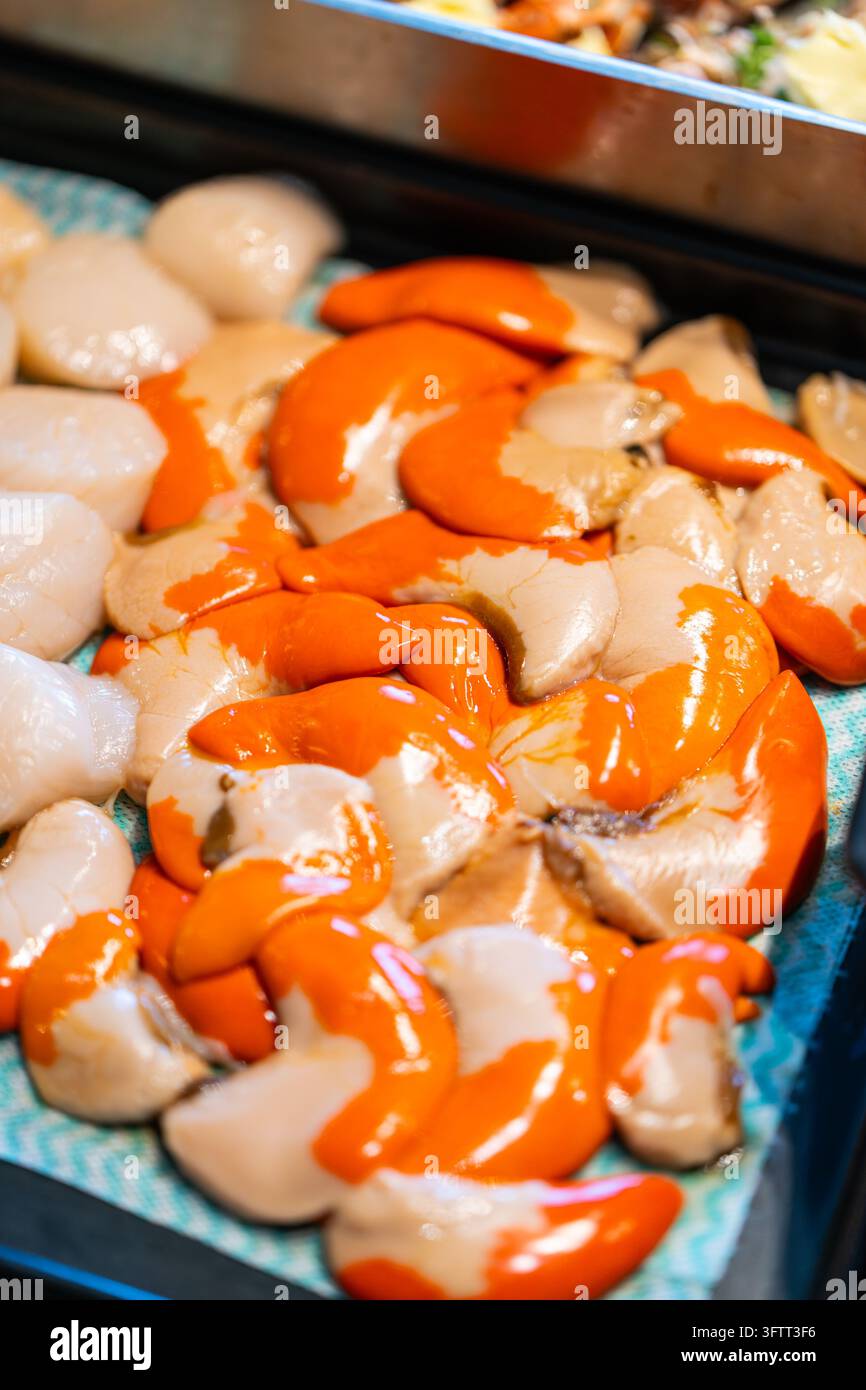 Capasante reali crude fresche con roe arancione brillante, o corallo, disposte su carta da cucina in un vassoio, con aragosta imburrata sfocata sullo sfondo Foto Stock