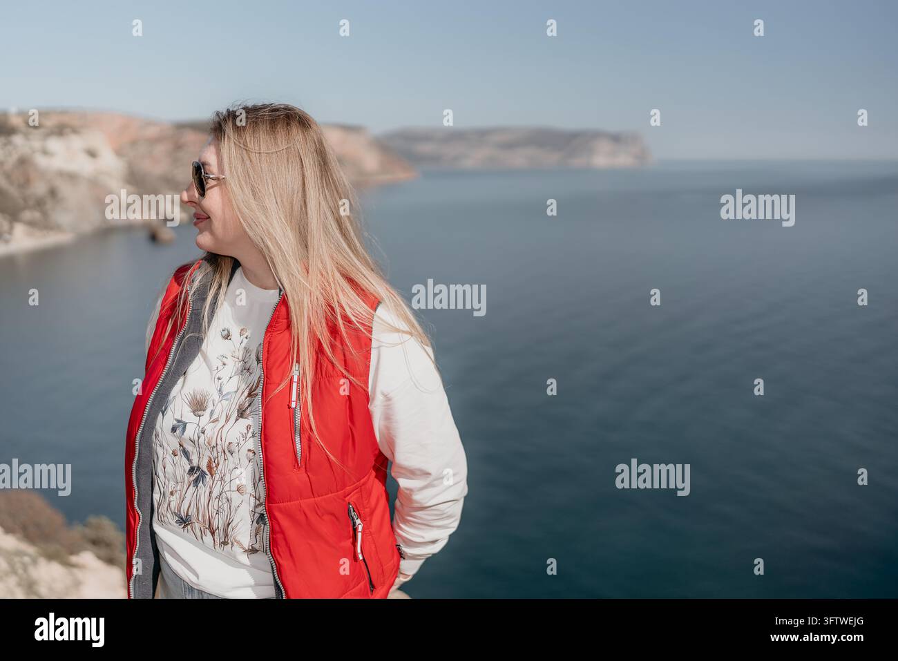 Donna viaggio costiero, signora bionda con un giubbotto rosso e occhiali da sole guarda il mare calmo da un alto dirupo durante una vacanza soleggiata Foto Stock