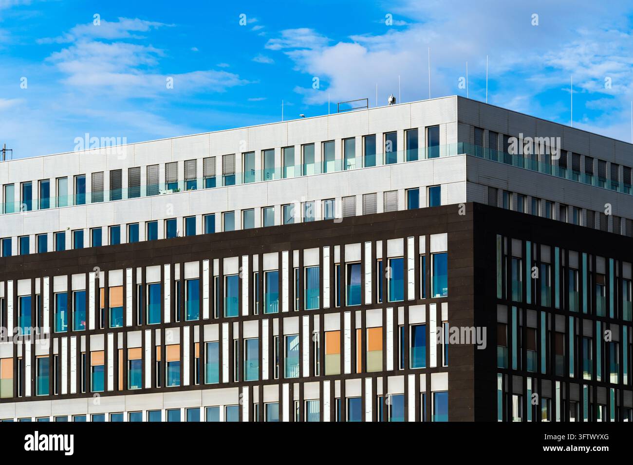 Edificio moderno d'ufficio con sezioni di facciata contrastanti bianche e scure, finestre rettangolari ripetute e accenti arancioni. La luce del sole evidenzia le Foto Stock