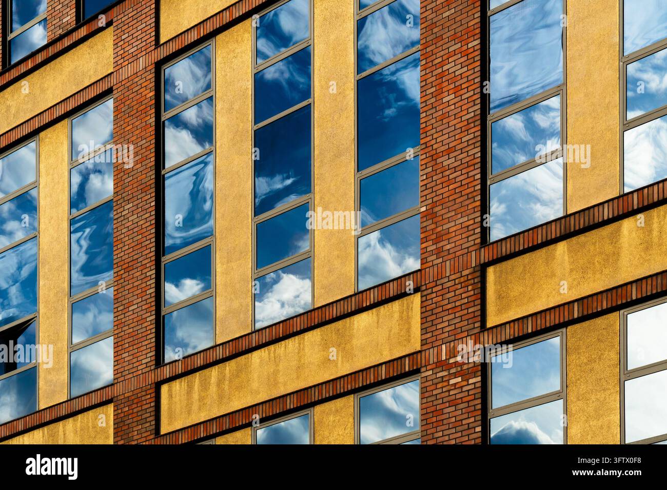 Finestre alte riflettono il cielo blu e le nuvole bianche turbinanti su una facciata gialla dell'edificio. Colonne di mattoni rossi e fasce orizzontali incorniciano la parete ruvida Foto Stock