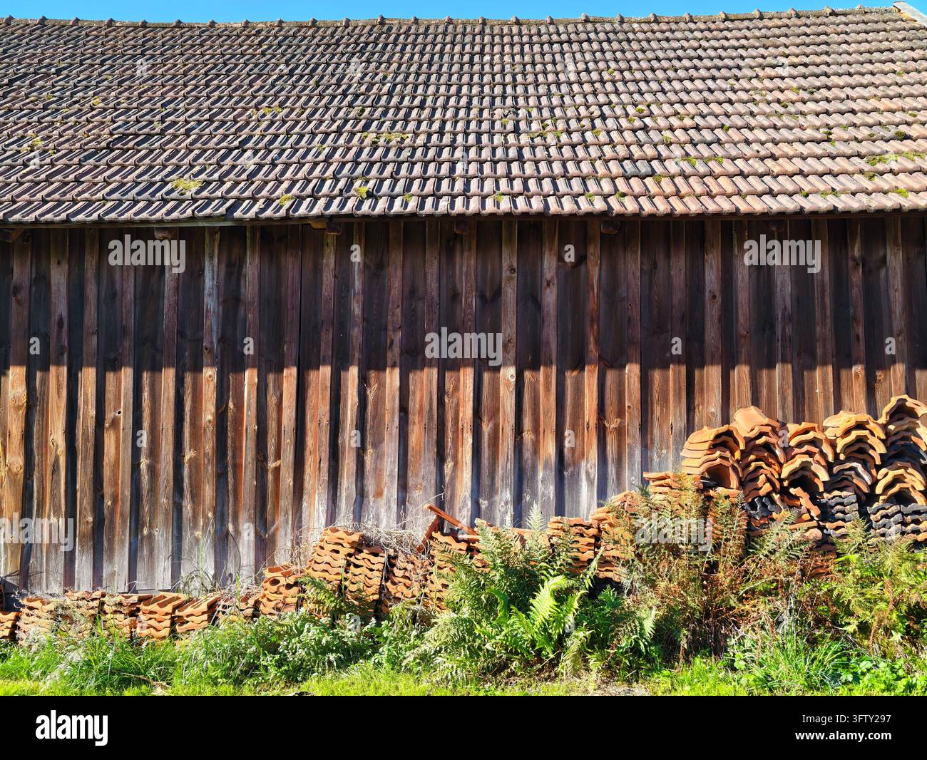 Stalla di legno stagionato con tetto di tegole e tegole d'argilla impilate in un giardino rurale Foto Stock