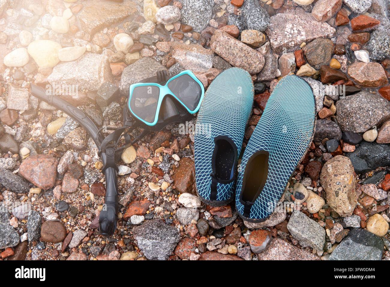 Maschera da snorkeling turchese, boccaglio e scarpe da acqua appoggiati su ciottoli e pietre multicolori bagnati con una luce soffusa che attraversa la scena. Foto Stock