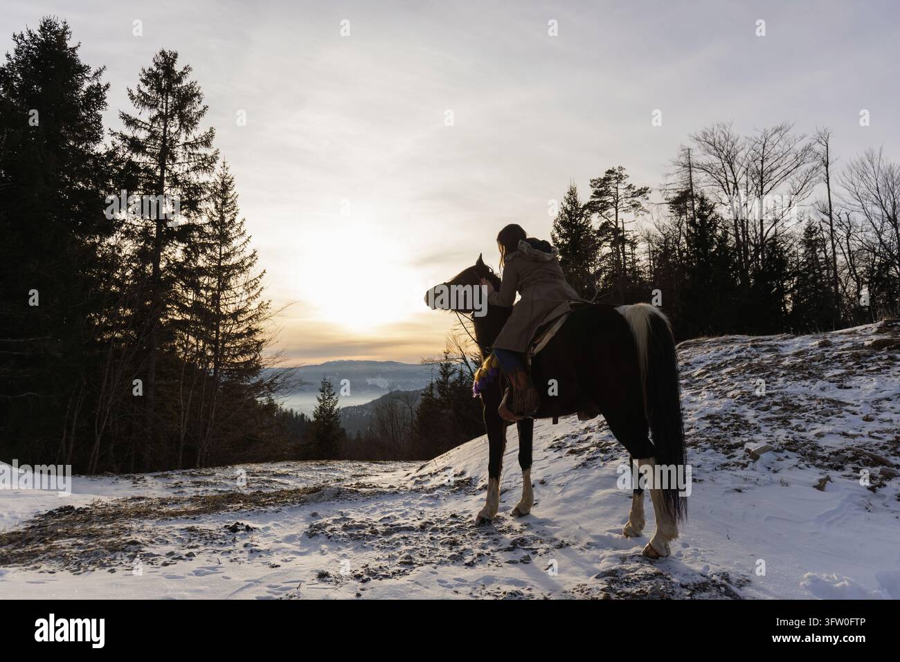 Guardando il tramonto da cavallo, una donna che godersi un momento di pace circondata da panorami montani e natura invernale. Foto Stock