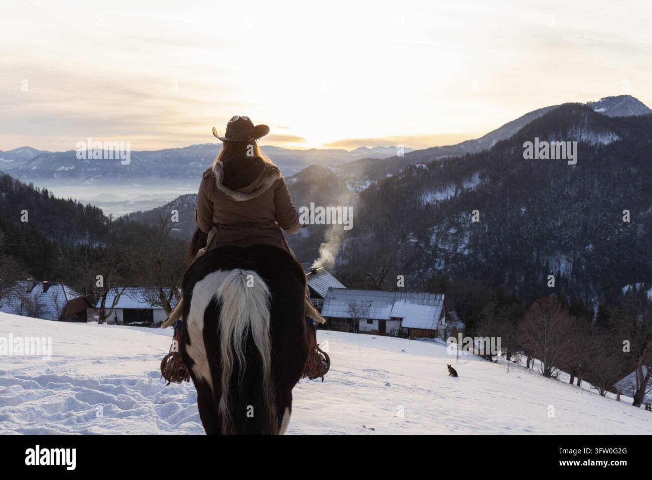 Donna a cavallo attraverso un paesaggio rurale innevato, che si gode una vacanza invernale attiva tra le montagne e la campagna tranquilla. Foto Stock