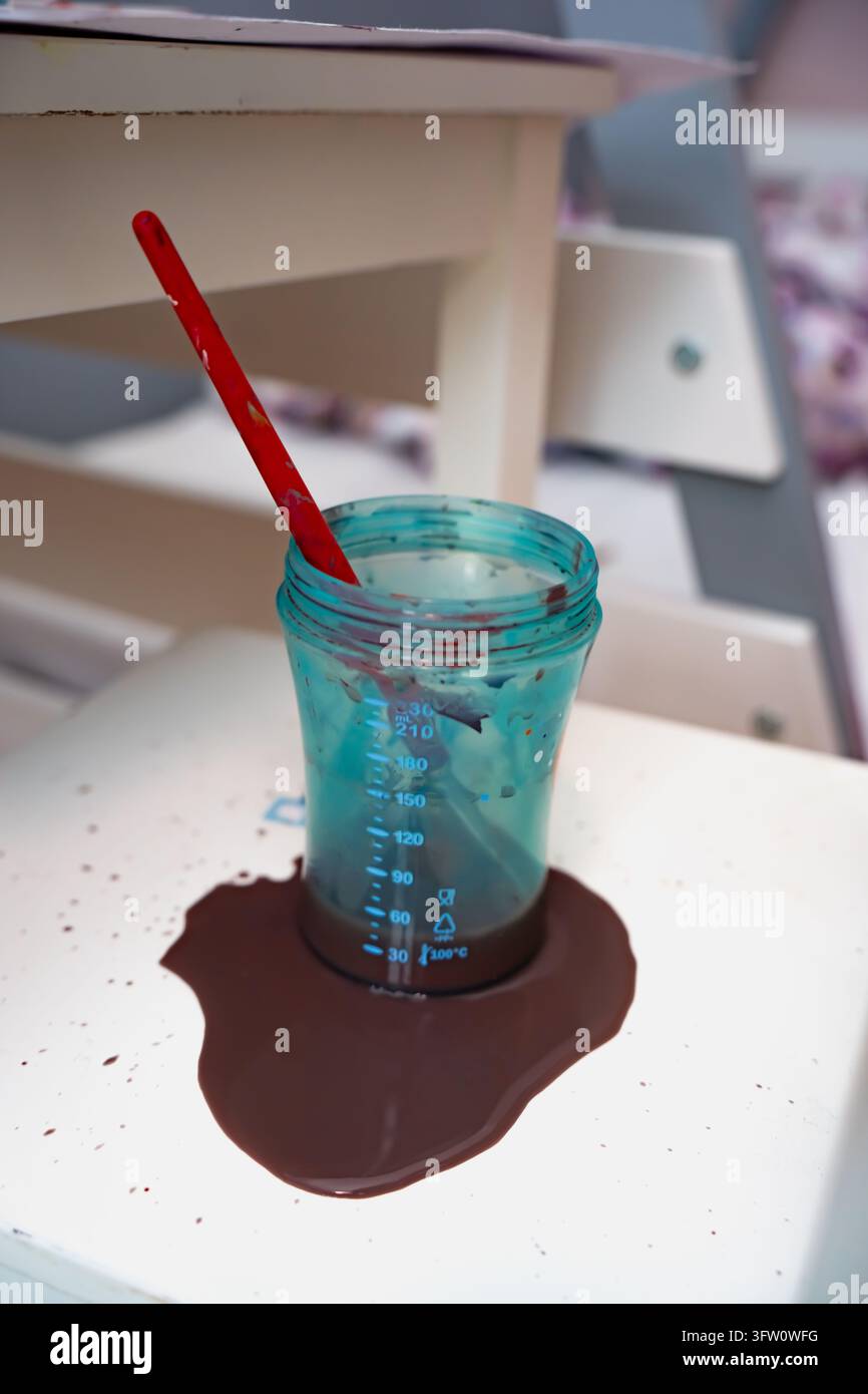 Un pennello si trova in una tazza di plastica graduata circondata da liquido versato su un banco da lavoro, illustrando la manipolazione degli strumenti per la pittura e le esigenze di pulizia presso un Foto Stock