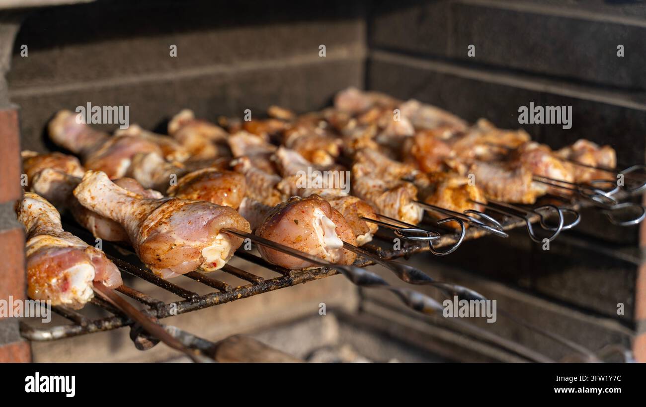 Primo piano di cosce di pollo crude condite infilate su spiedini metallici e disposte su una griglia all'interno di una grigliaia a carbone. Foto Stock