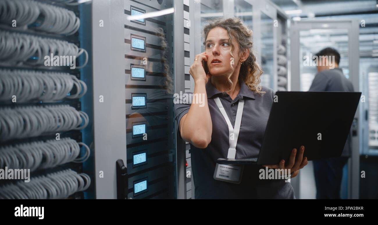 Specialista IT donna controlla lo stato del server su laptop e schermi LED del rack mentre parla al telefono, con un collega maschio che effettua la manutenzione di un altro armadio dietro di lei in un moderno data center, mostrando il lavoro di squadra. Foto Stock