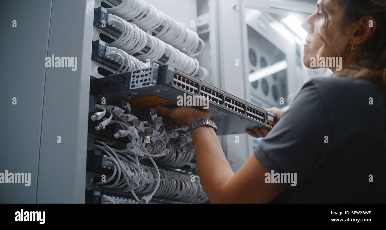 Tecnico di rete femminile installa hardware di switch attivo in rack di server, completando la sostituzione e l'aggiornamento delle apparecchiature di rete per un'infrastruttura aziendale affidabile, connettività e servizio dati. Foto Stock