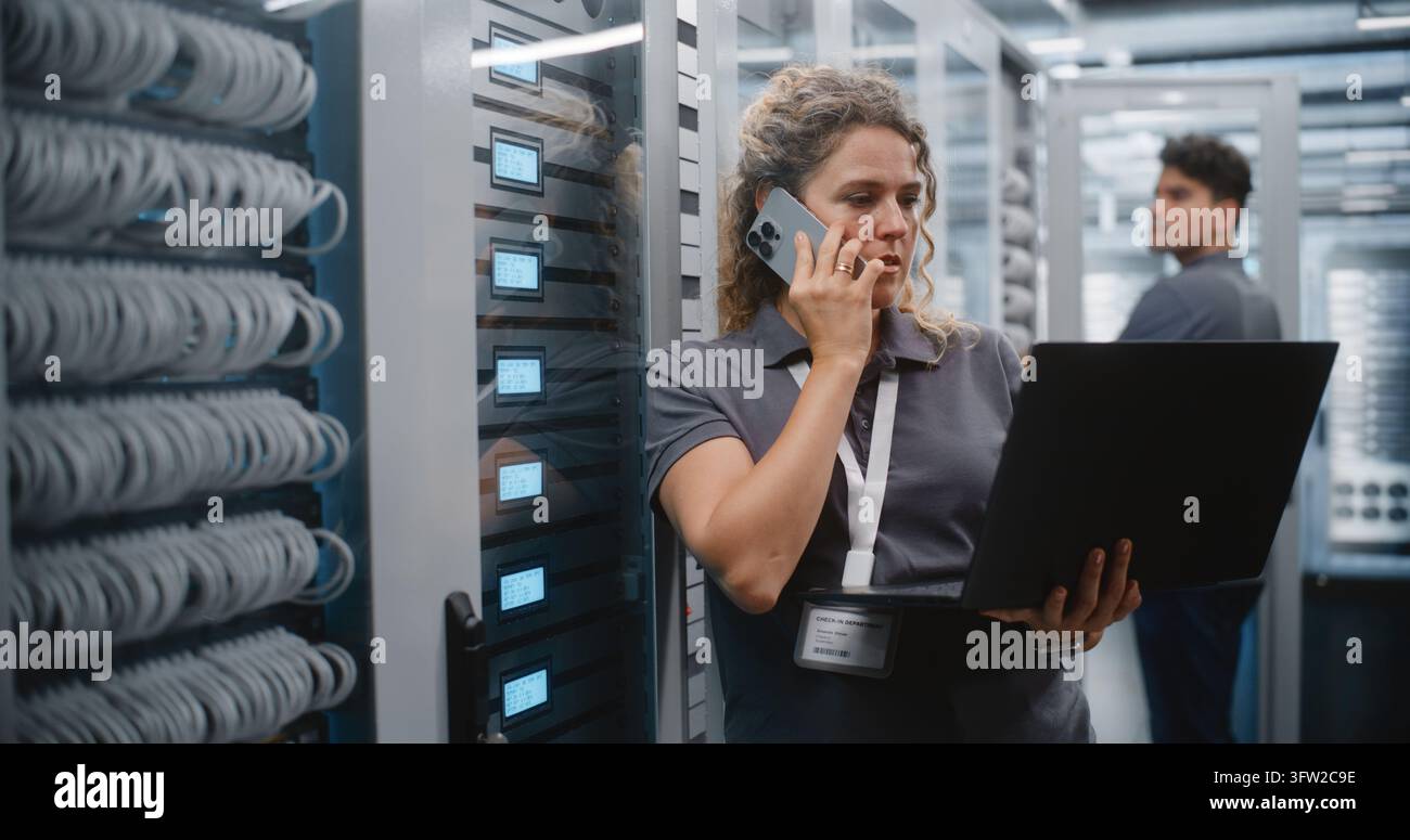 Specialista IT donna controlla lo stato del server su laptop e schermi LED del rack mentre parla al telefono, con un collega maschio che effettua la manutenzione di un altro armadio dietro di lei in un moderno data center, mostrando il lavoro di squadra. Foto Stock