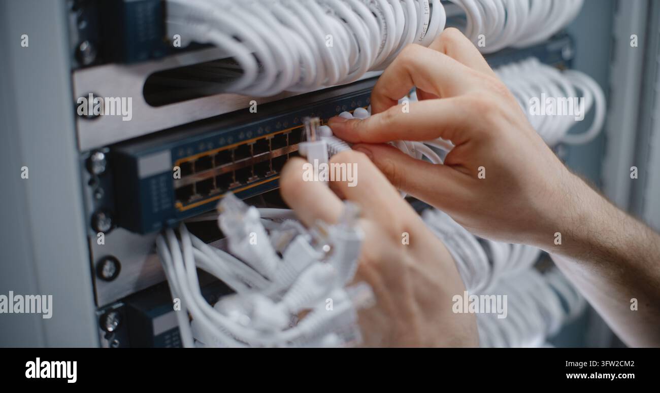 Primo piano di una mano di uno specialista maschile che esegue la manutenzione del cablaggio del rack, collegando cavi di collegamento e organizzando fasci di cavi tra il pannello di patch e lo switch per un'infrastruttura pulita e una connettività LAN stabile. Foto Stock