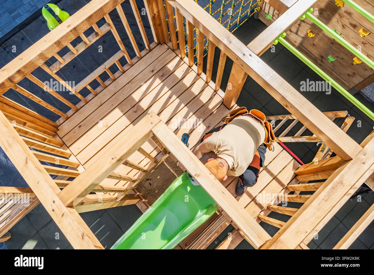 Un operaio assembla una torre da gioco in legno con ringhiere e uno scivolo verde, vista dall'alto attraverso la struttura in legno. Foto Stock
