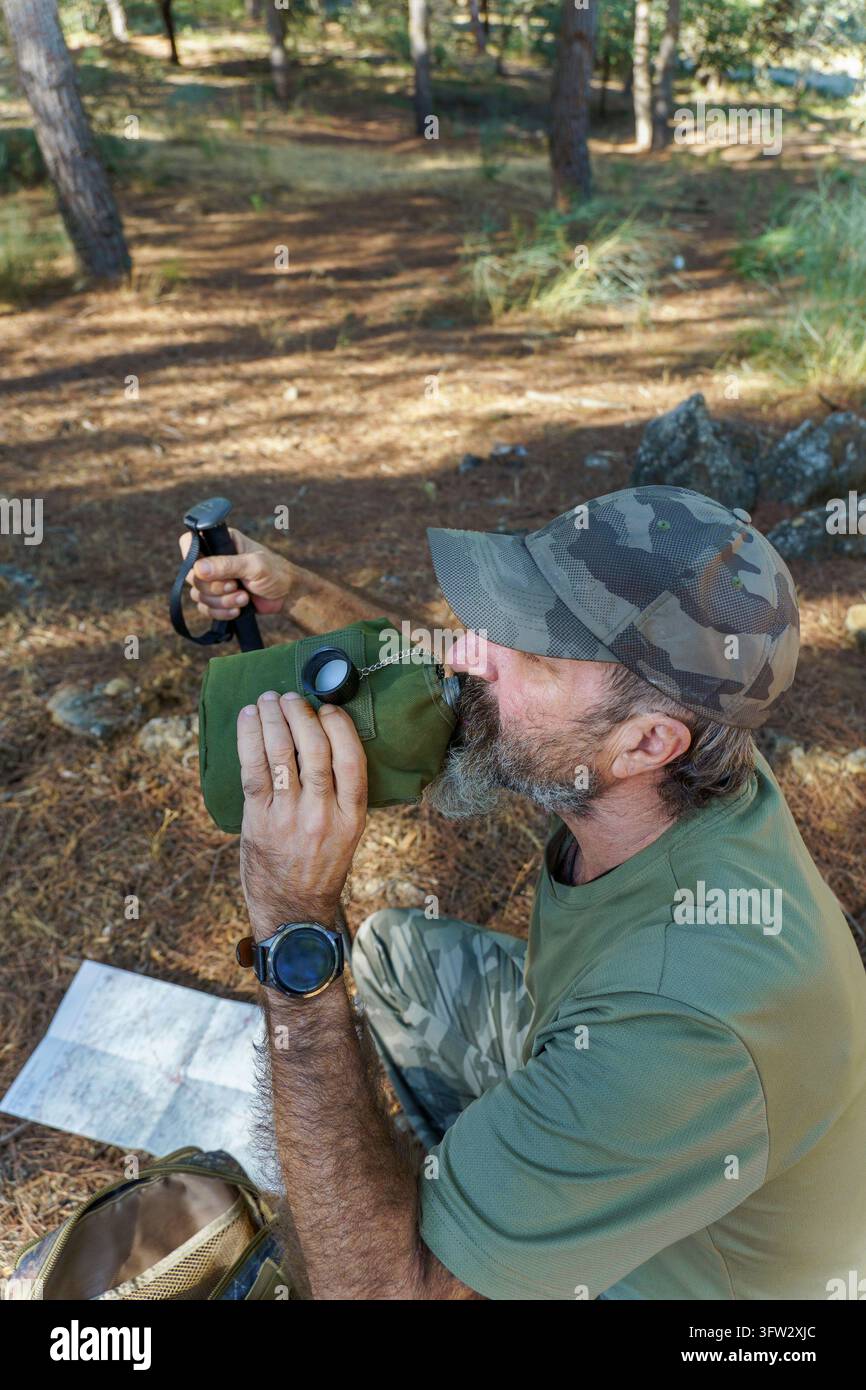 Uomo barbuto con cappello e abiti mimetici che fa una pausa per bere acqua dalla sua borraccia durante un'escursione in una foresta Foto Stock