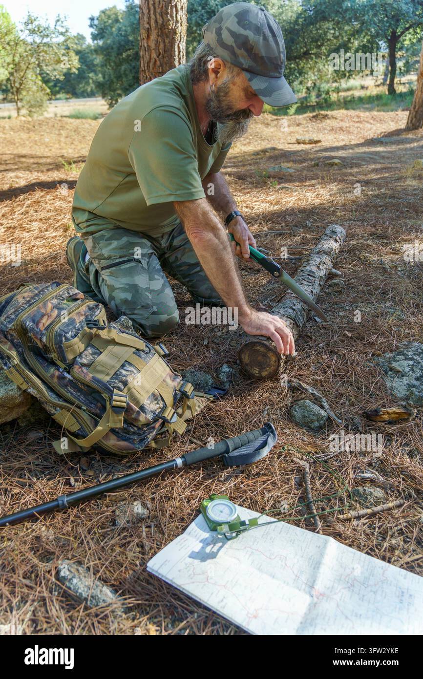 Uomo barbuto in abiti mimetici che segna un tronco con una sega a mano. Z Zaino, bastone da trekking, mappa e bussola giacevano a terra Foto Stock