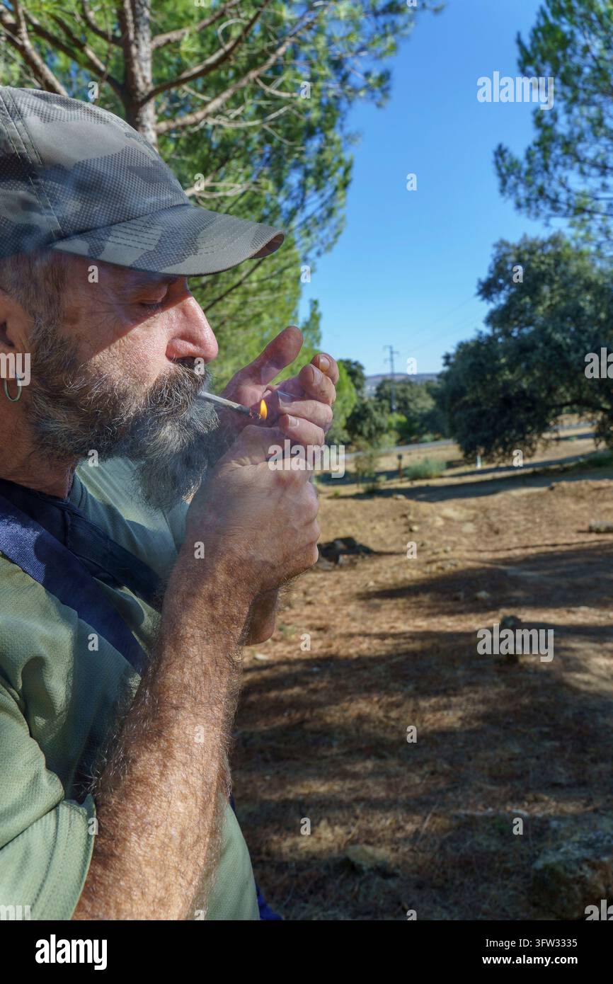 Uomo barbuto con cappello mimetico che accende una sigaretta arrotolata in una foresta soleggiata, vista di lato, momento di relax all'aperto Foto Stock