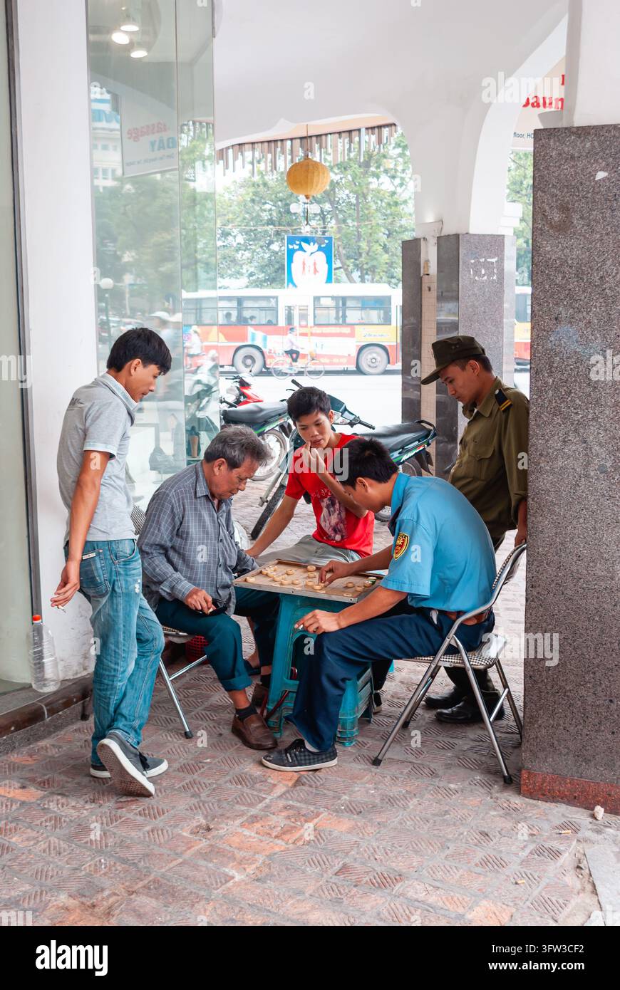 HANOI, VIETNAM - 16 LUGLIO 2011: I residenti locali si radunano sotto un edificio commerciale nel centro di Hanoi per giocare a un gioco da tavolo tradizionale mentre il traffico p Foto Stock