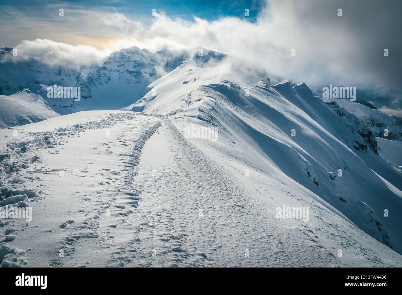 Una vista panoramica spettacolare di una cresta montuosa innevata con nuvole basse che fluttuano sopra le cime frastagliate e impronte di passi incise nella polvere profonda e ghiacciata Foto Stock