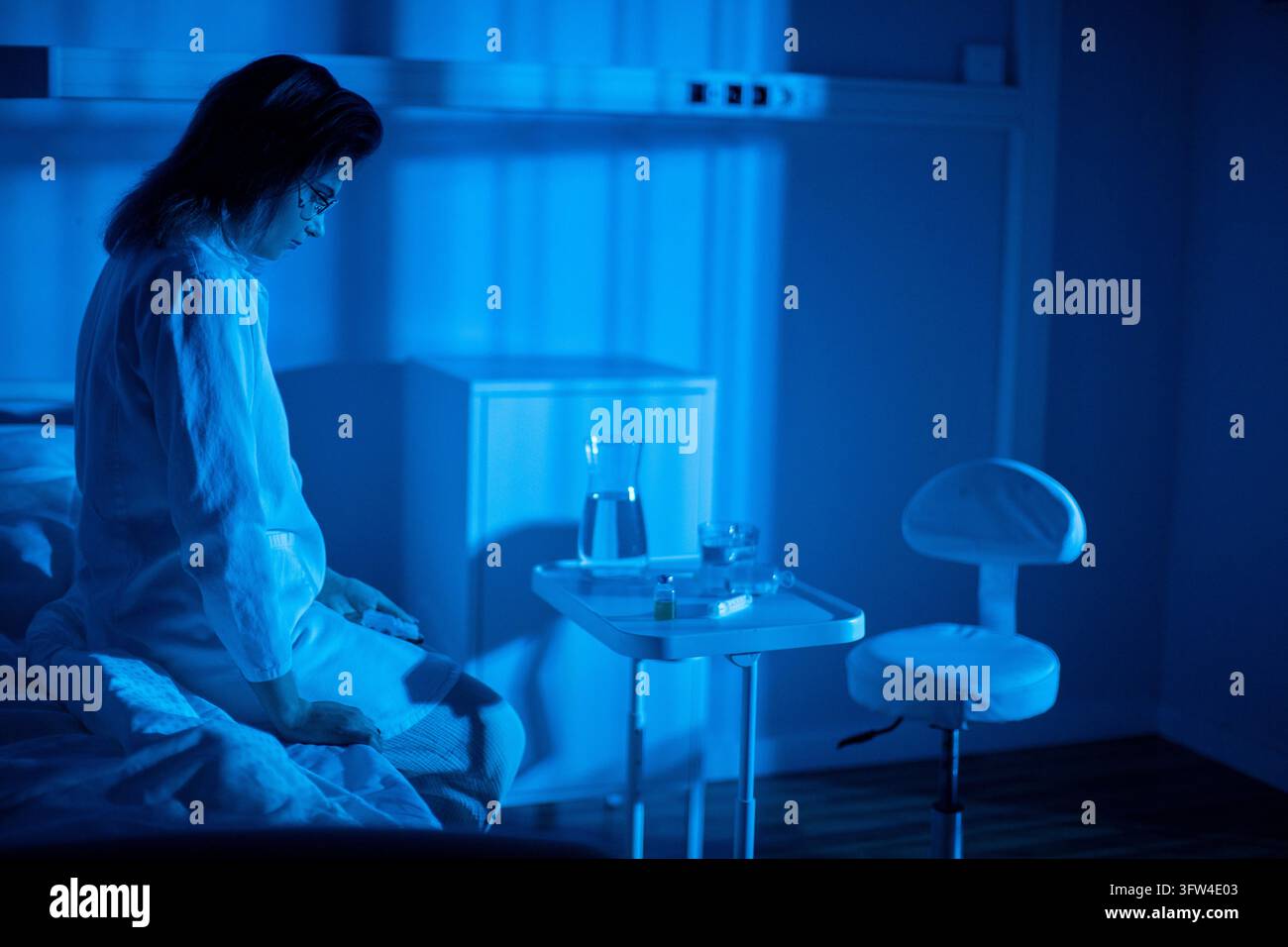 Il paziente è seduto sul letto vicino alla finestra, pigiama spiegazzato, lampada che proietta una luce blu soffusa, mani giunte mentre fissa le luci lontane, momento tranquillo di guarigione Foto Stock