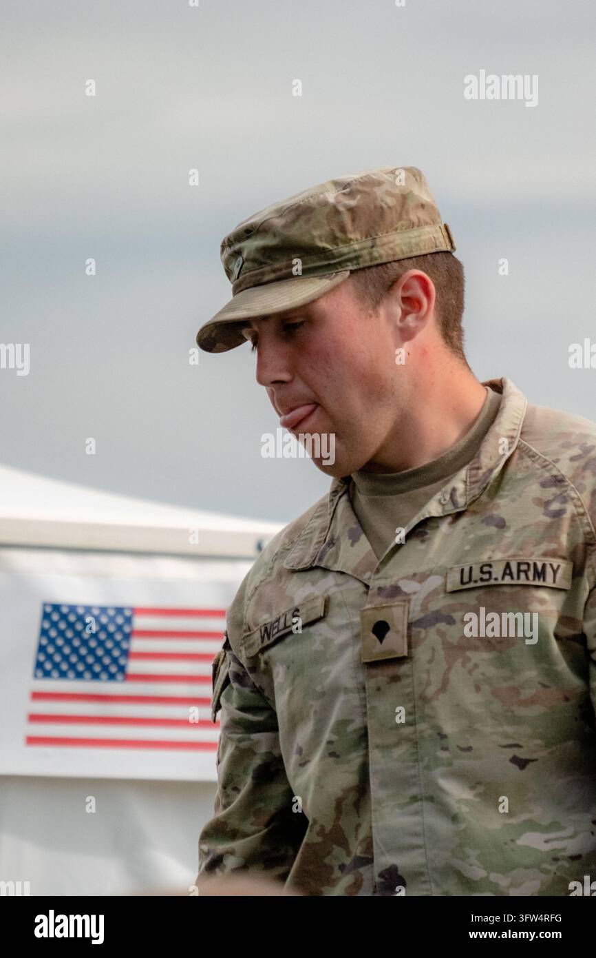 Giovane soldato con berretto da pattuglia mimetico e uniforme in piedi vicino a uno striscione della bandiera americana, che guarda verso il basso con contemplazione Foto Stock