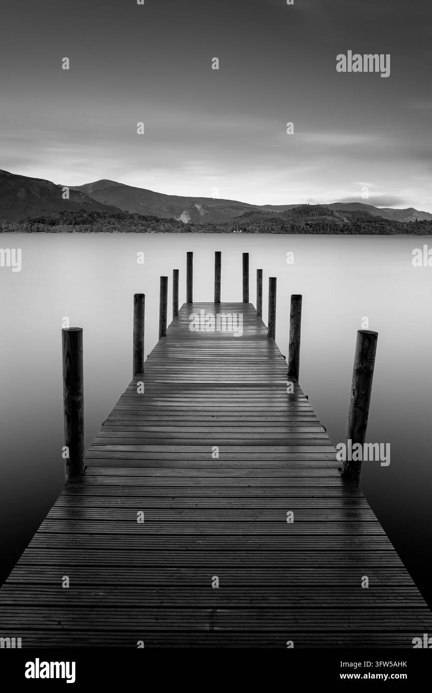 Inghilterra, Cumbria, Distretto dei Laghi, Derwentwater. Un molo di legno eroso si protende in acque calme, con montagne boschive e un cielo dolcemente nuvoloso Foto Stock