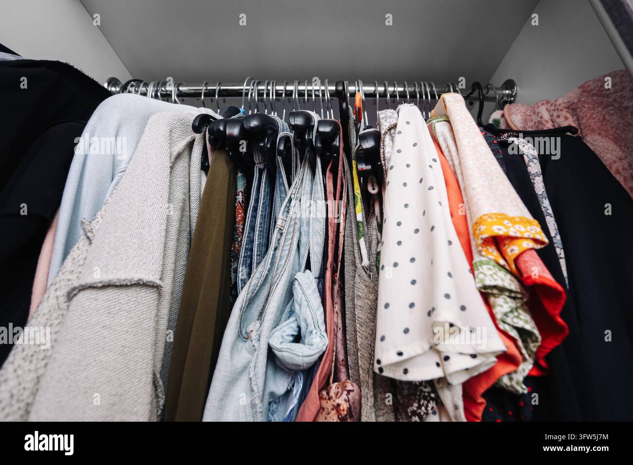 Guardaroba affollata con vari abiti appesi su una rastrelliera all'interno di un armadio bianco. Conservazione domestica di abiti con camicie, giacche e vestiti in spazio limitato. Foto Stock