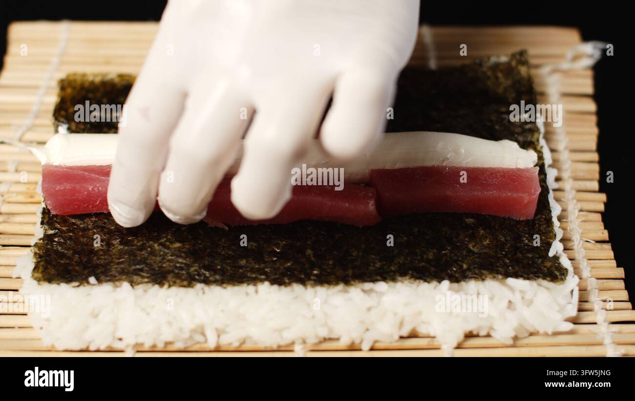 Le mani guantate dello chef preparano un rotolo di sushi al tonno fresco su un materassino di bambù con riso, alghe e crema di formaggio Foto Stock