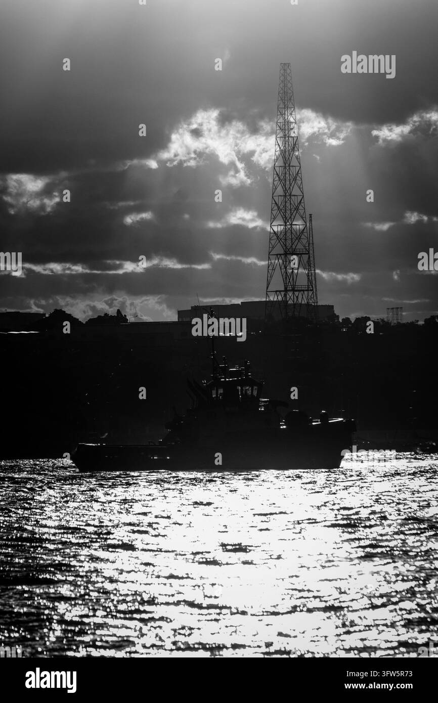 Silhouette di un rimorchiatore al tramonto con torre industriale e raggi di sole drammatici sull'acqua Foto Stock