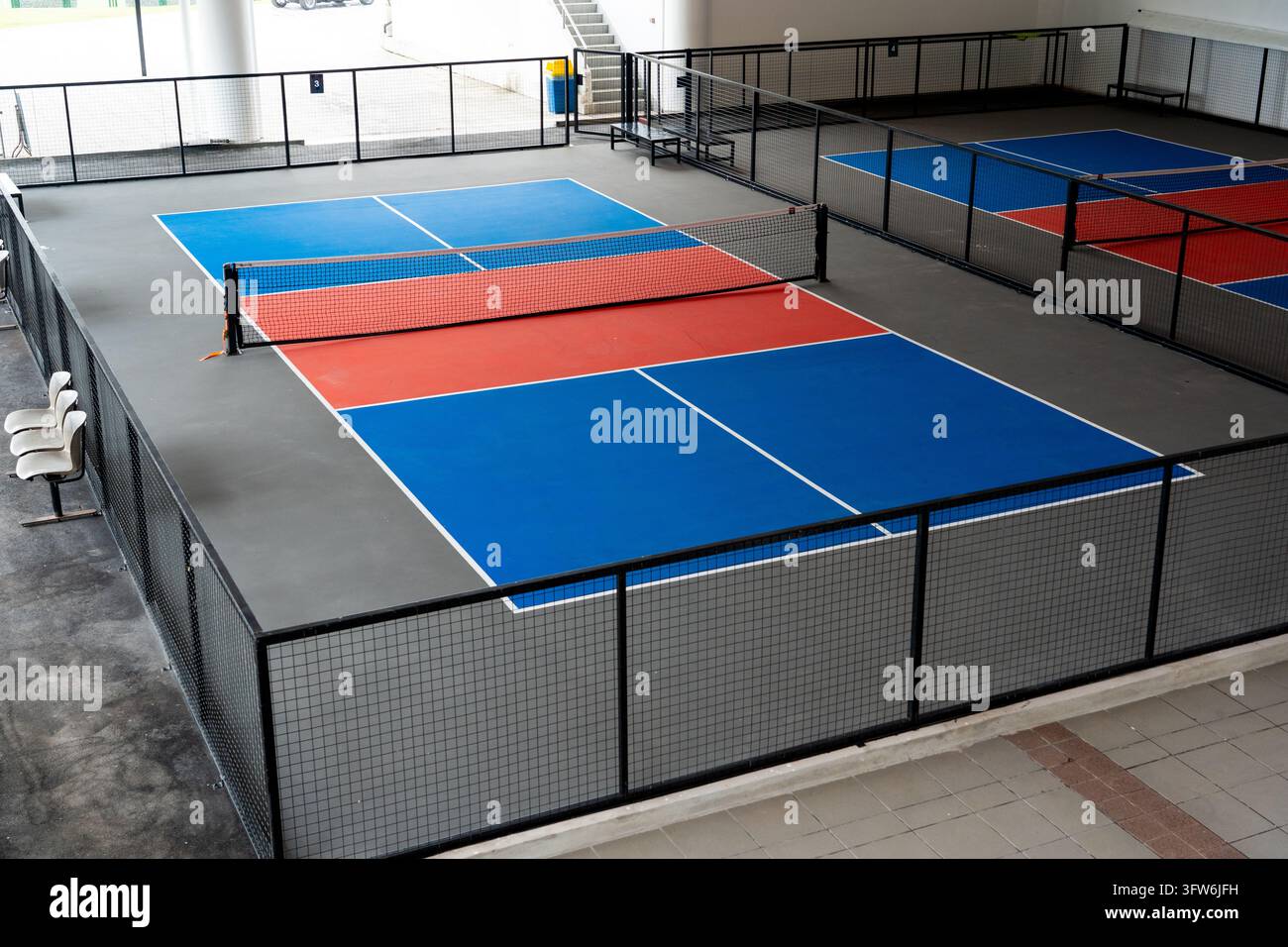 Campi da pickleball indoor moderni vuoti con superfici dure dipinte di blu e rosso, reti nere, recinzioni di sicurezza in rete metallica e area seduta in un Foto Stock