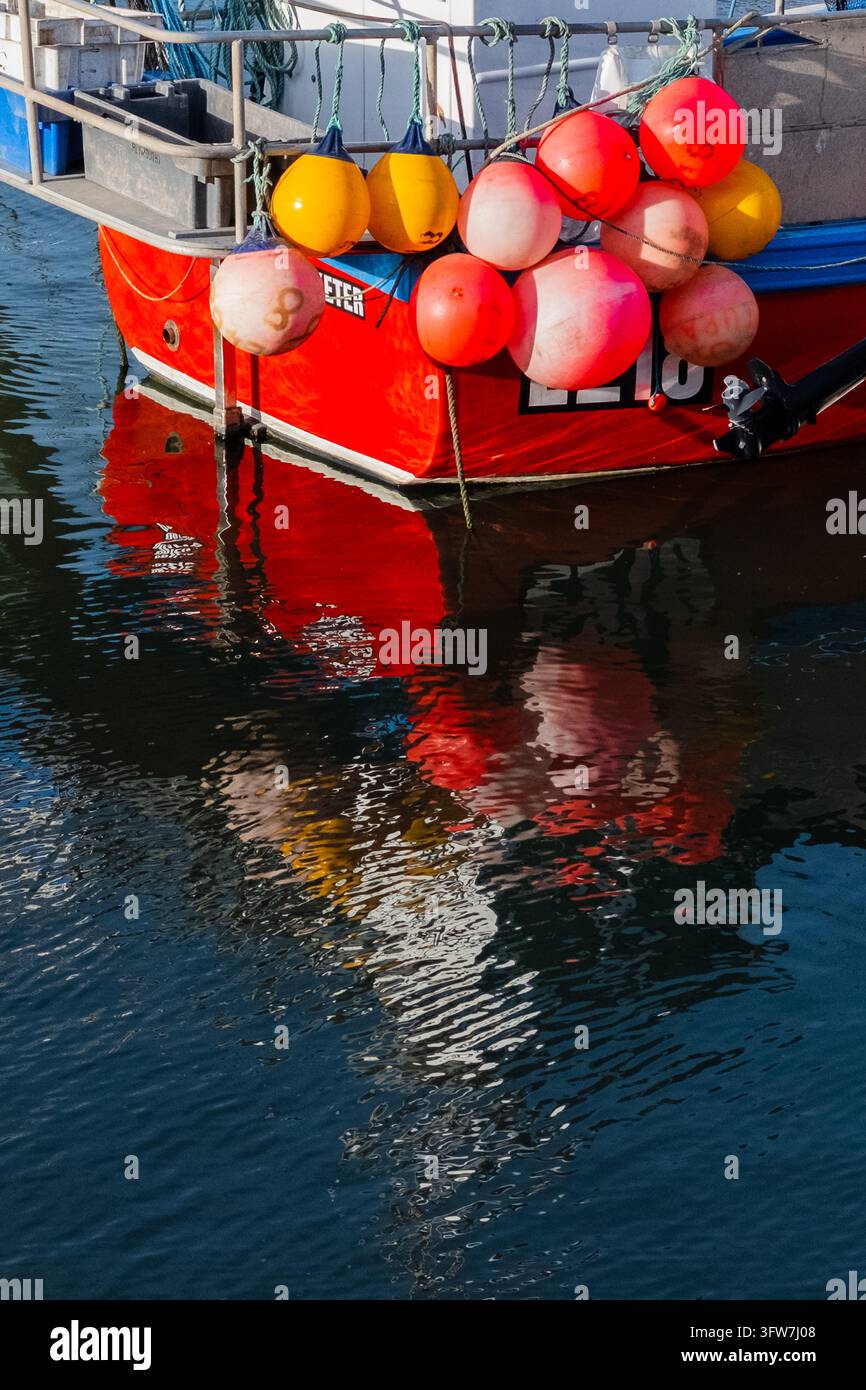 Poppa di un peschereccio rosso acceso con boe colorate che si riflettono nelle increspature dell'acqua calma del porto di West Bay Harbour, Dorset, Inghilterra, Regno Unito Foto Stock