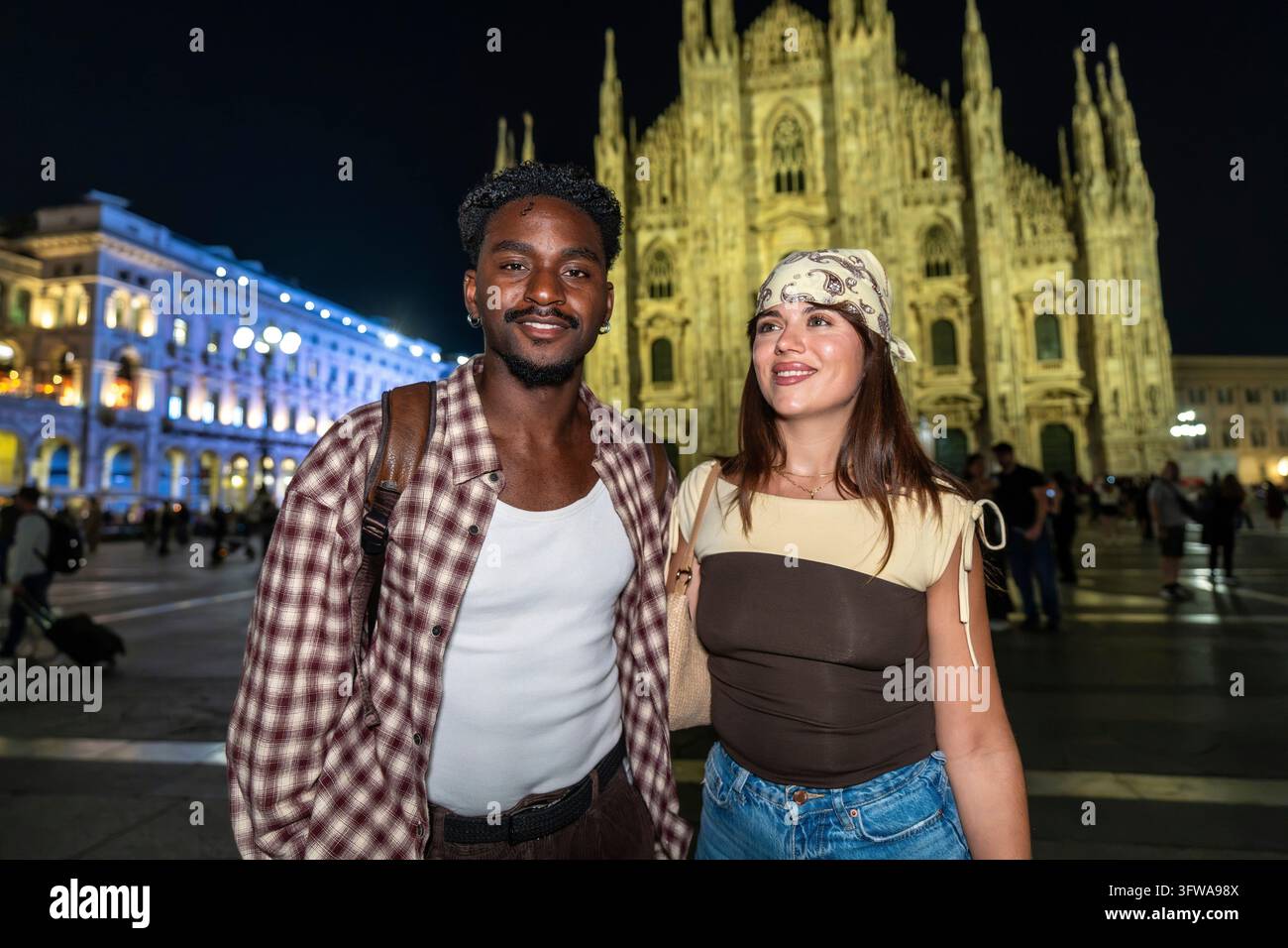 Giovane coppia multirazziale che sorride alla telecamera, in piedi davanti al duomo illuminato a Milano, Italia, vivendo il turismo urbano europeo Foto Stock
