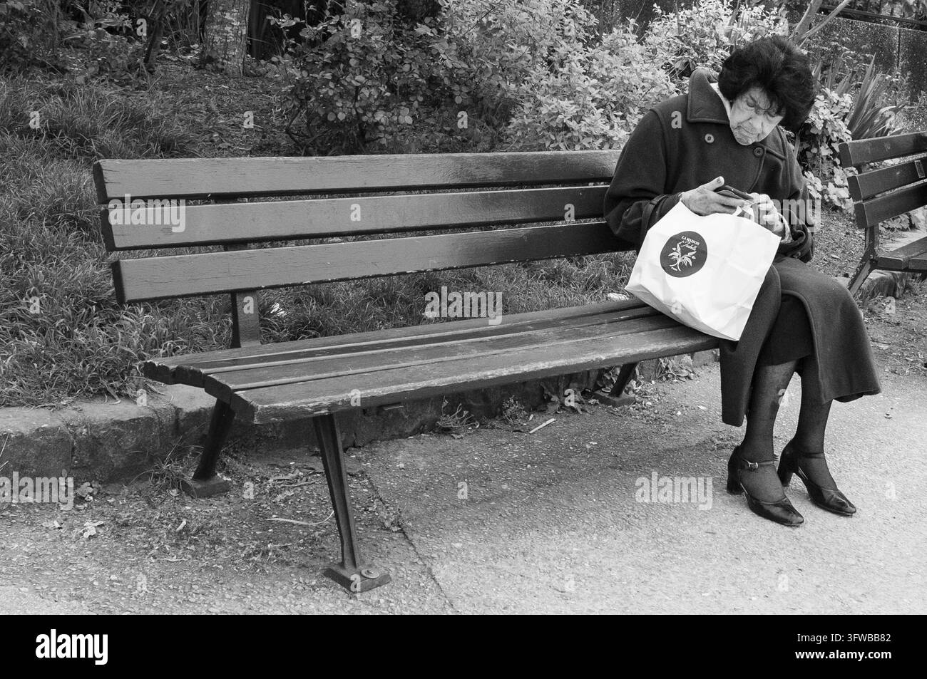 DONNA ANZIANA SU PANCHINA DEL PARCO CON TELEFONO, REPORTAGE DELLA DOMENICA SULLA RIVA SINISTRA DELLA SEINE, PARIGI, FRANCIA, 2026: Un'elegante donna anziana in un cappotto blu è seduta da sola su una panchina verde nel parco, tiene una borsa della spesa e guarda il suo smartphone. INFO: La riva sinistra a est di Notre-Dame unisce eredità accademica con romanticismo fluviale. Passeggiando davanti alle università storiche, ai tranquilli giardini botanici e al moderno Istituto del Mondo Arabo si rivela un angolo vivace e meno turistico di Parigi, pulsante di anima intellettuale. Foto Stock