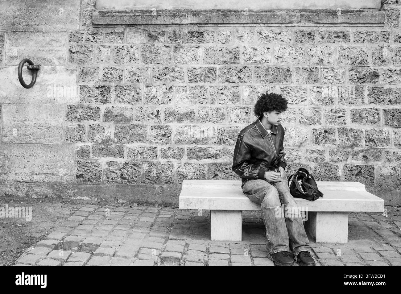 GIOVANE UOMO SEDUTO SU UNA PANCHINA, SUA RICORDAZIONE DELLA RIVA SINISTRA DELLA DOMENICA, PARIGI, FRANCIA, 2026: Un giovane elegante con una grande acconciatura afro, che indossa una giacca di pelle nera e jeans, è seduto da solo su una moderna panchina di cemento davanti a un vecchio muro di pietra. INFO: La riva sinistra a est di Notre-Dame unisce eredità accademica con romanticismo fluviale. Passeggiando davanti a università storiche, tranquilli giardini botanici e il moderno Istituto del Mondo Arabo si rivela un angolo vivace e meno turistico di Parigi, pulsante di anima intellettuale. Foto Stock