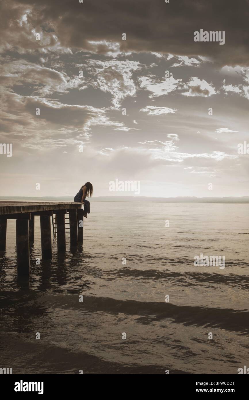 Miniatura di una donna seduta da sola su un molo sul mare, che simboleggia solitudine, introspezione e riflessione emotiva Foto Stock