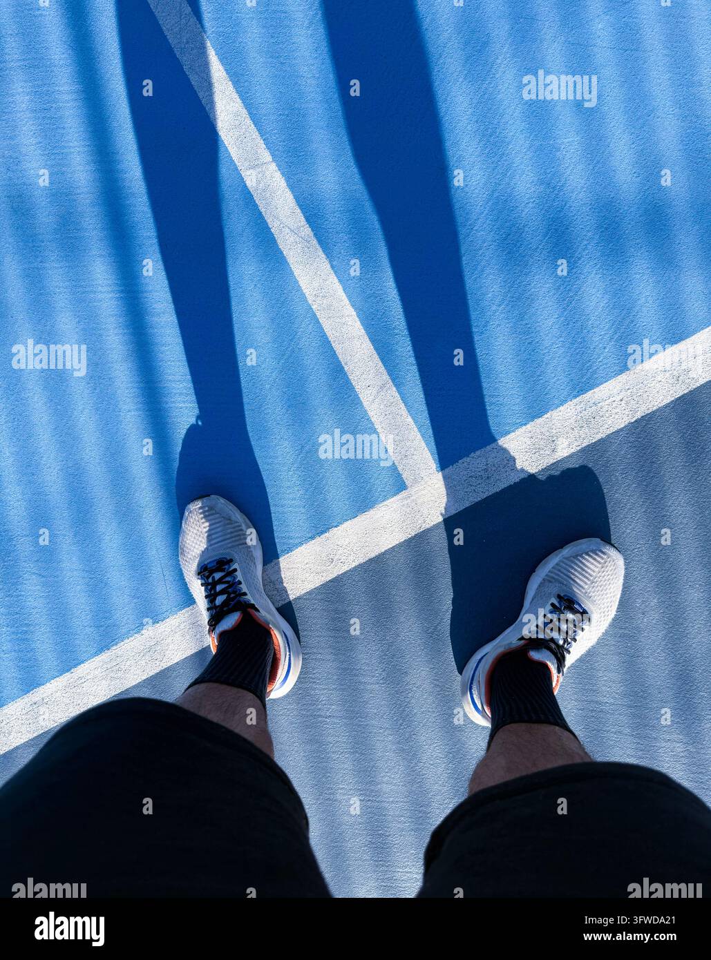 Persona in piedi su un campo blu con linee bianche e ombre di recinzione, indossa scarpe da ginnastica, pronta per l'azione Foto Stock