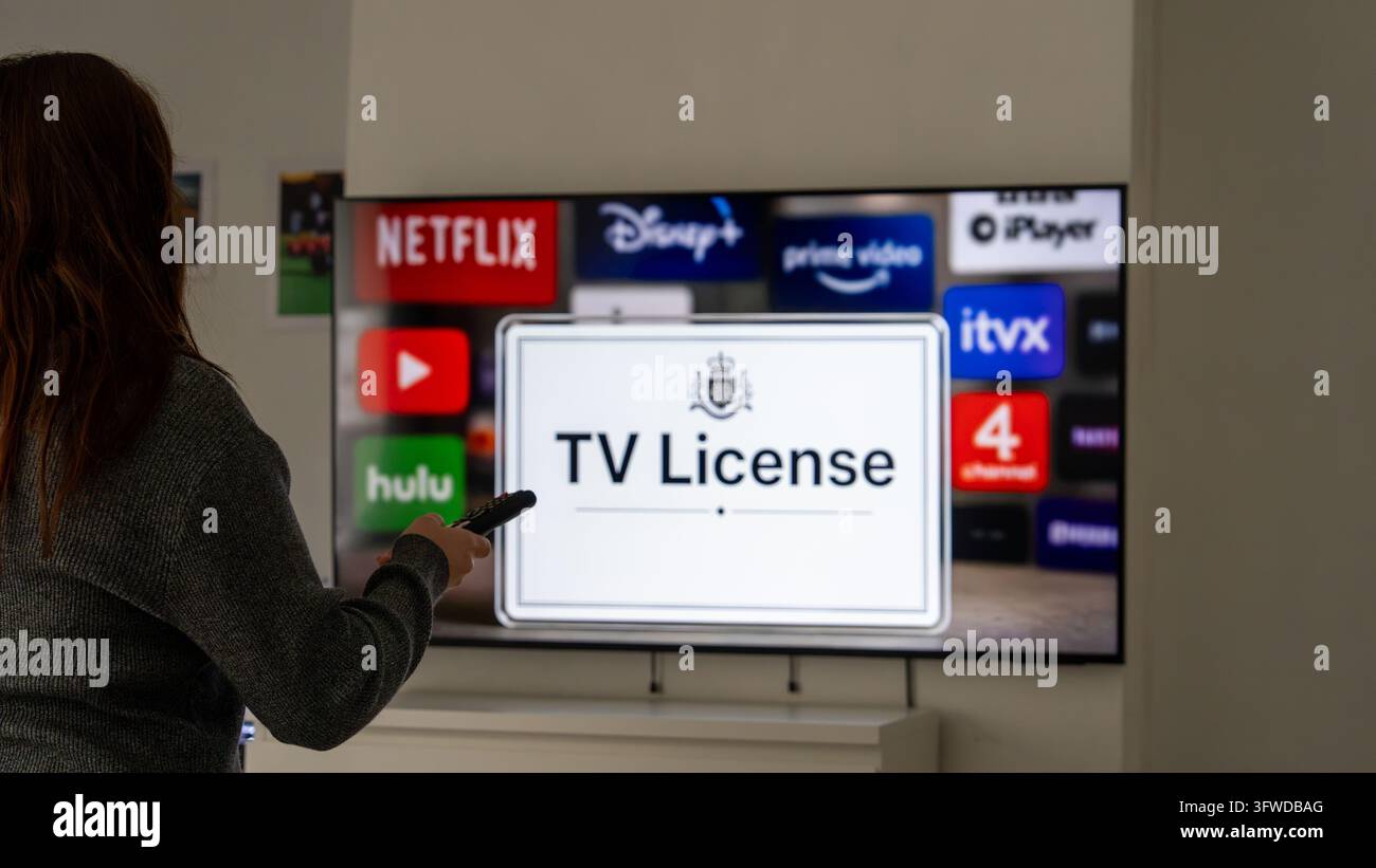Donna indica un televisore intelligente con una remoto, mostrando un avviso di licenza TV, con i loghi delle app di streaming popolari sullo sfondo a casa. Foto Stock