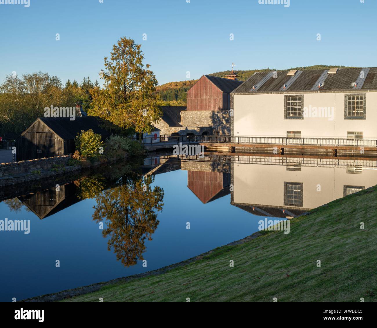 Gli edifici e gli alberi della distilleria Glenfiddich si riflettono sulla superficie del suo stagno di raffreddamento a Dufftown, Moray, Scozia. Foto Stock