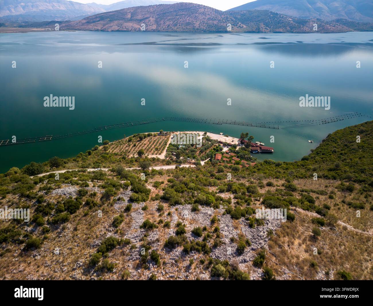 Le infrastrutture per l'allevamento di cozze formano file parallele su un lago protetto. Il sito di acquacoltura include edifici sulla riva e un molo, incorniciati da un terreno roccioso Foto Stock