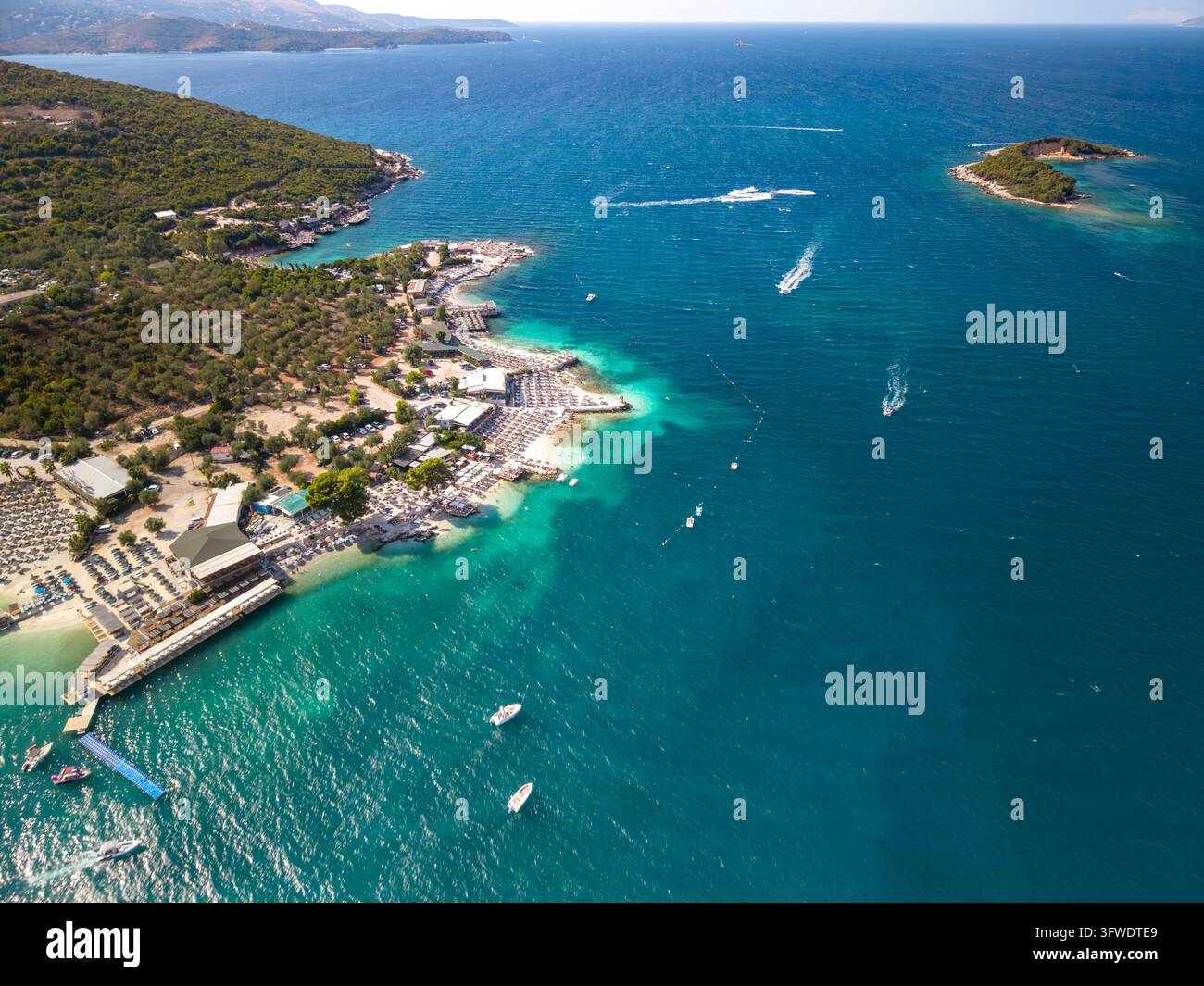 La costa di Ksamil in Albania confina con un mare turchese chiaro, con club di spiaggia e lettini che costeggiano la riva. Una vista aerea rivela una penisola boscosa, un piccolo offshore-i Foto Stock
