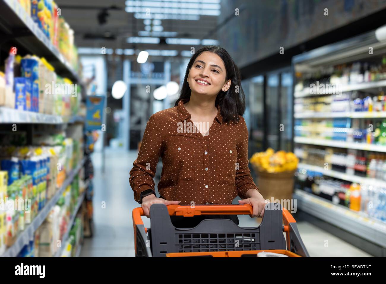 Giovane donna che spinge un carrello della spesa lungo un corridoio luminoso del supermercato, sorridendo e guardando in alto mentre seleziona prodotti freschi, godendosi un'atmosfera allegra e moder Foto Stock