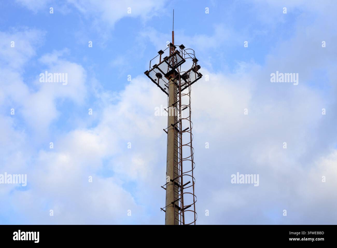 Alto palo di cemento con piattaforma metallica, scala e parafulmine si erge contro un cielo blu nuvoloso. Struttura industriale tecnica, torre di floodlight e Foto Stock