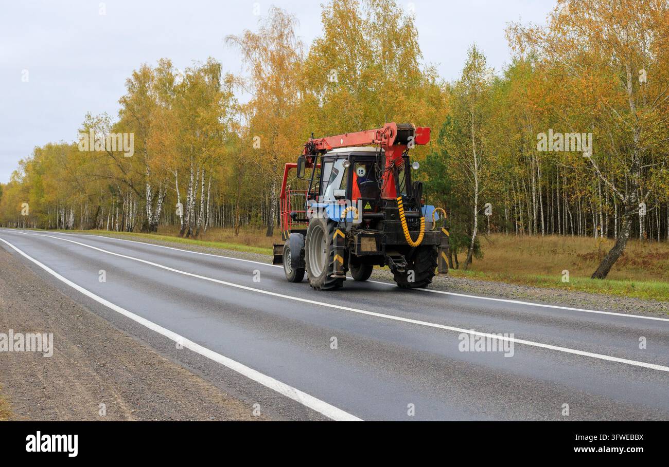 Trattore agricolo blu equipaggiato con braccio idraulico che procede lungo una strada asfaltata rurale accanto a una foresta autunnale. Meccanica utilitaria, servizio municipale Foto Stock