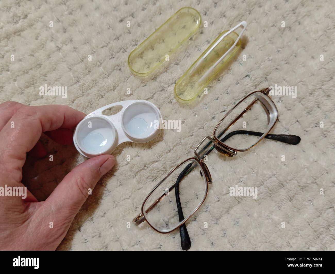 Una persona tiene una custodia per lenti a contatto mentre un paio di occhiali riposa su una superficie morbida a casa. La scena mostra oggetti utilizzati per la cura della vista. Foto Stock