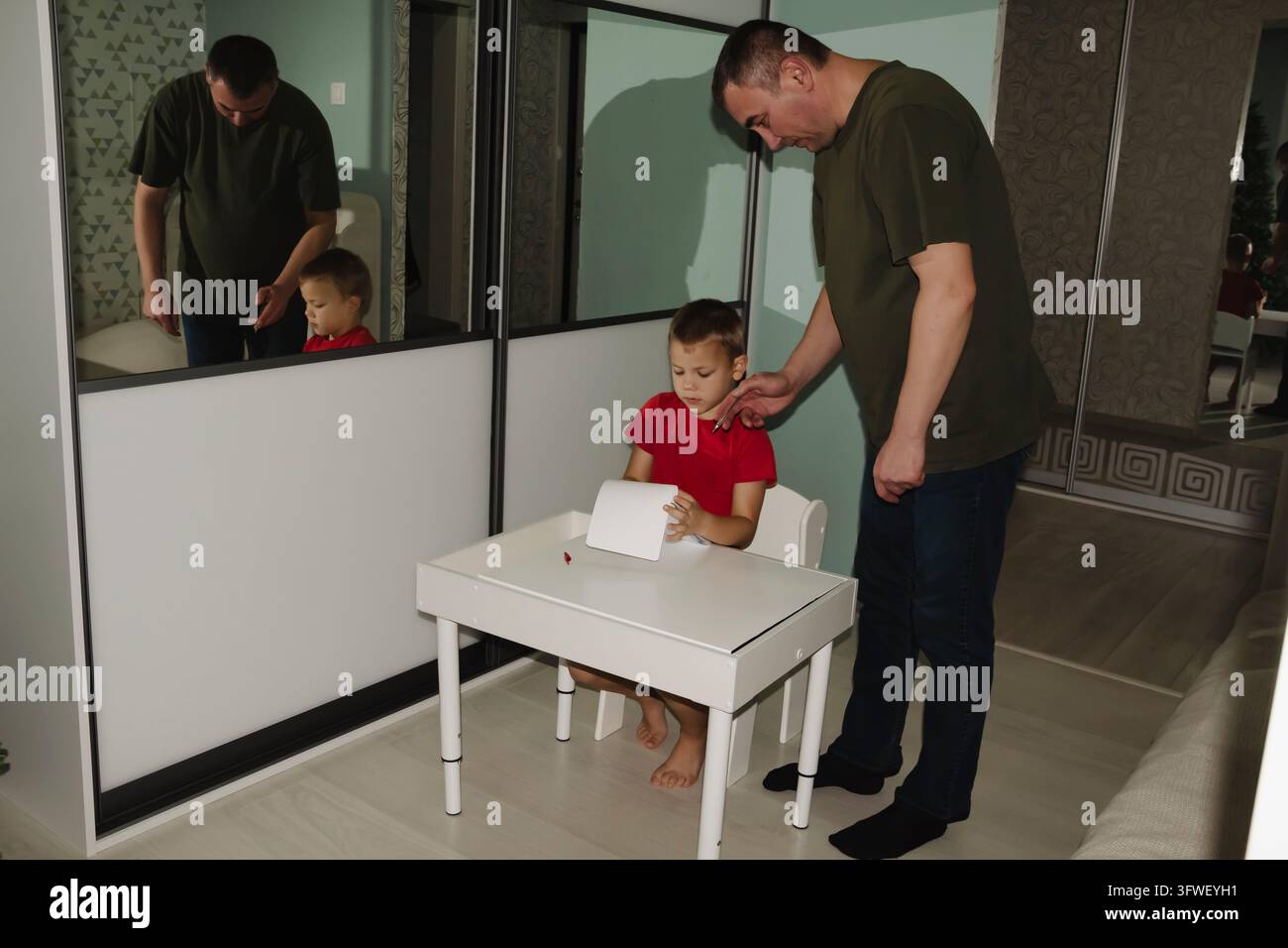 Un bambino torna da scuola indossando una maglietta rossa e si siede a un tavolo bianco per fare i compiti e i compiti per casa con l'aiuto di suo padre Foto Stock