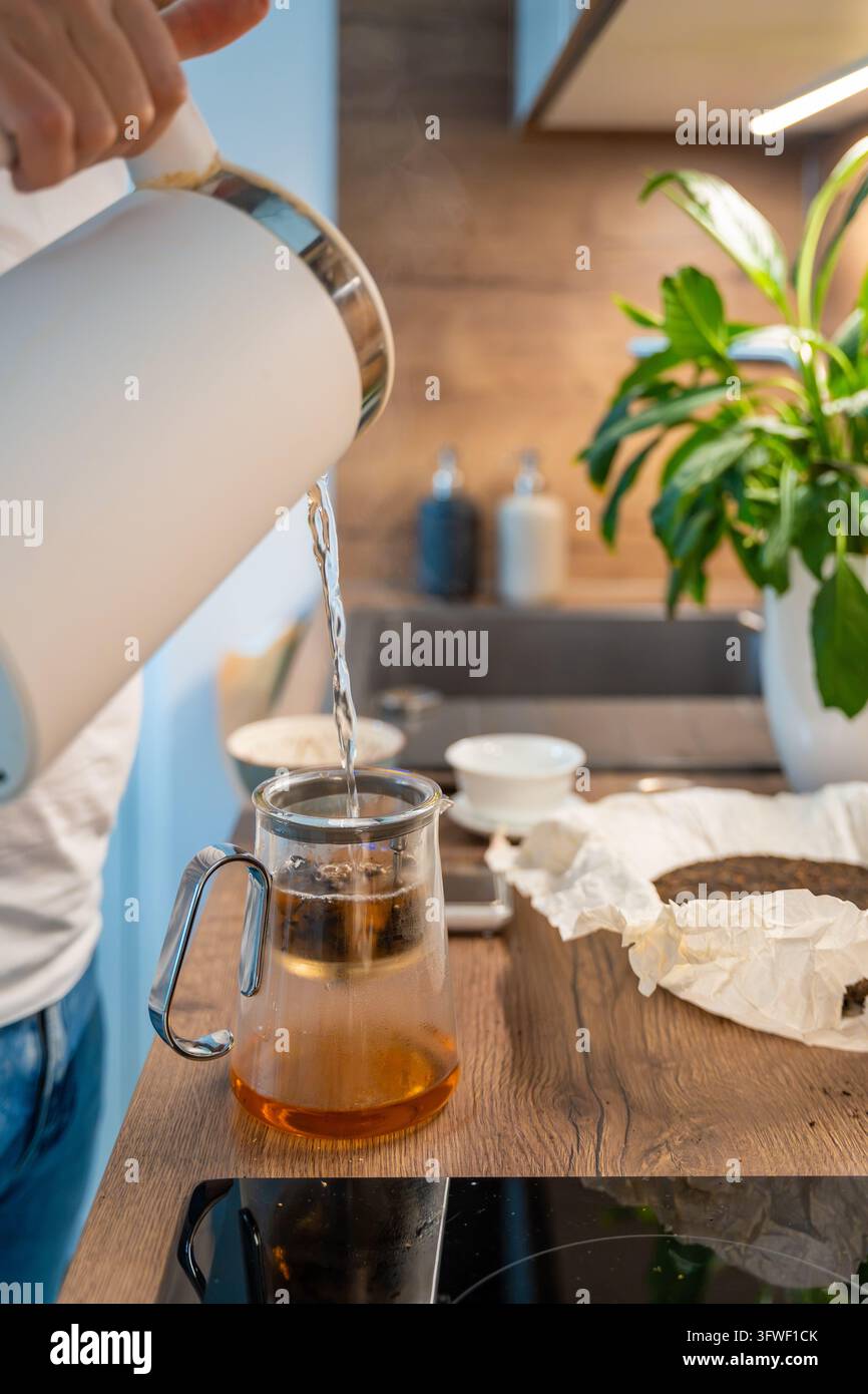 L'acqua scorre da un bollitore elettrico in un infusore da tè puer di vetro. Una breve infusione iniziale prepara le foglie per infusioni ripetute. Foto Stock
