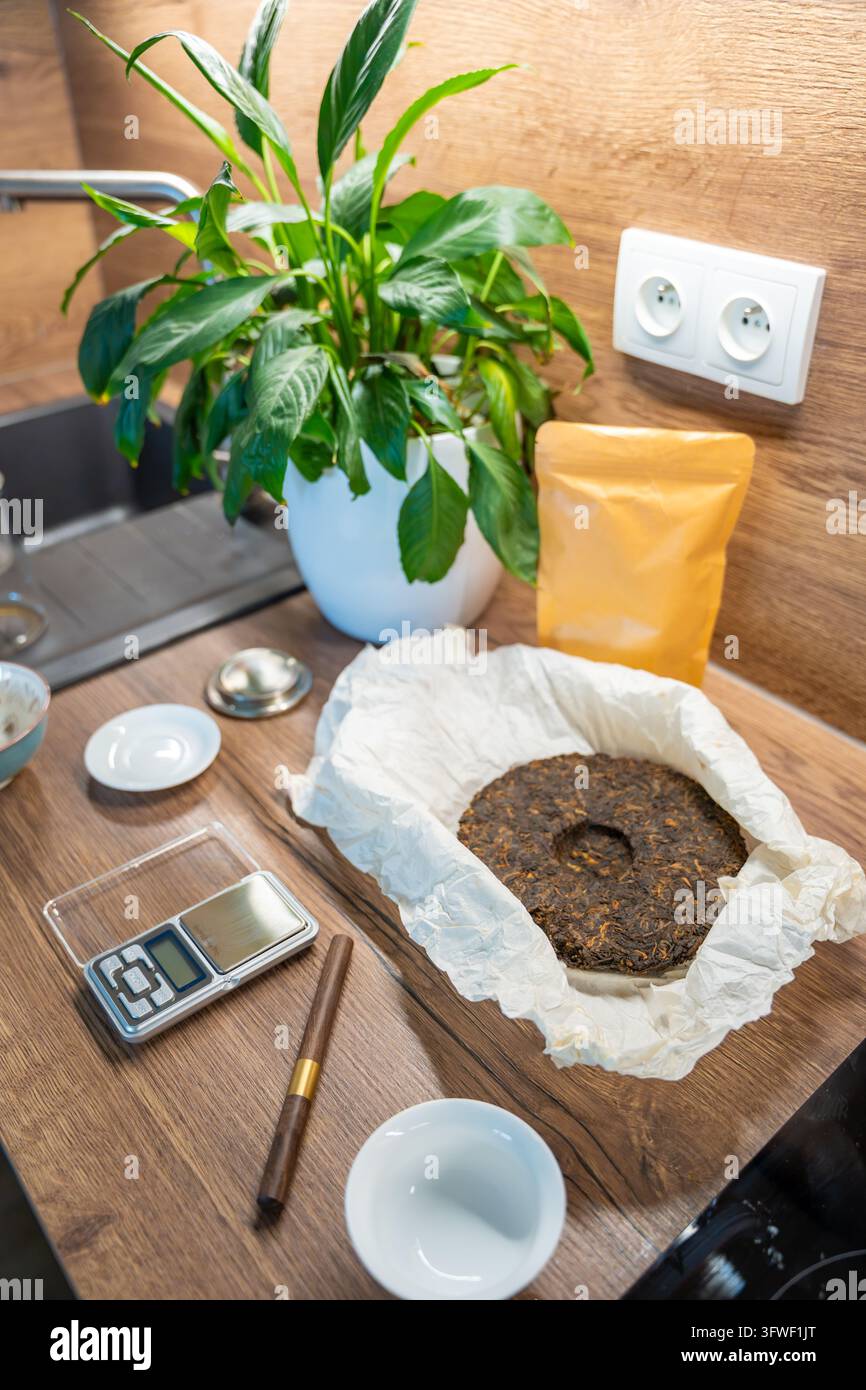Una torta di tè Puer compressa giace su carta accanto a una bilancia digitale e una pinza da tè. Misurare porzioni sciolte prepara il tè cinese per la preparazione domestica. Foto Stock