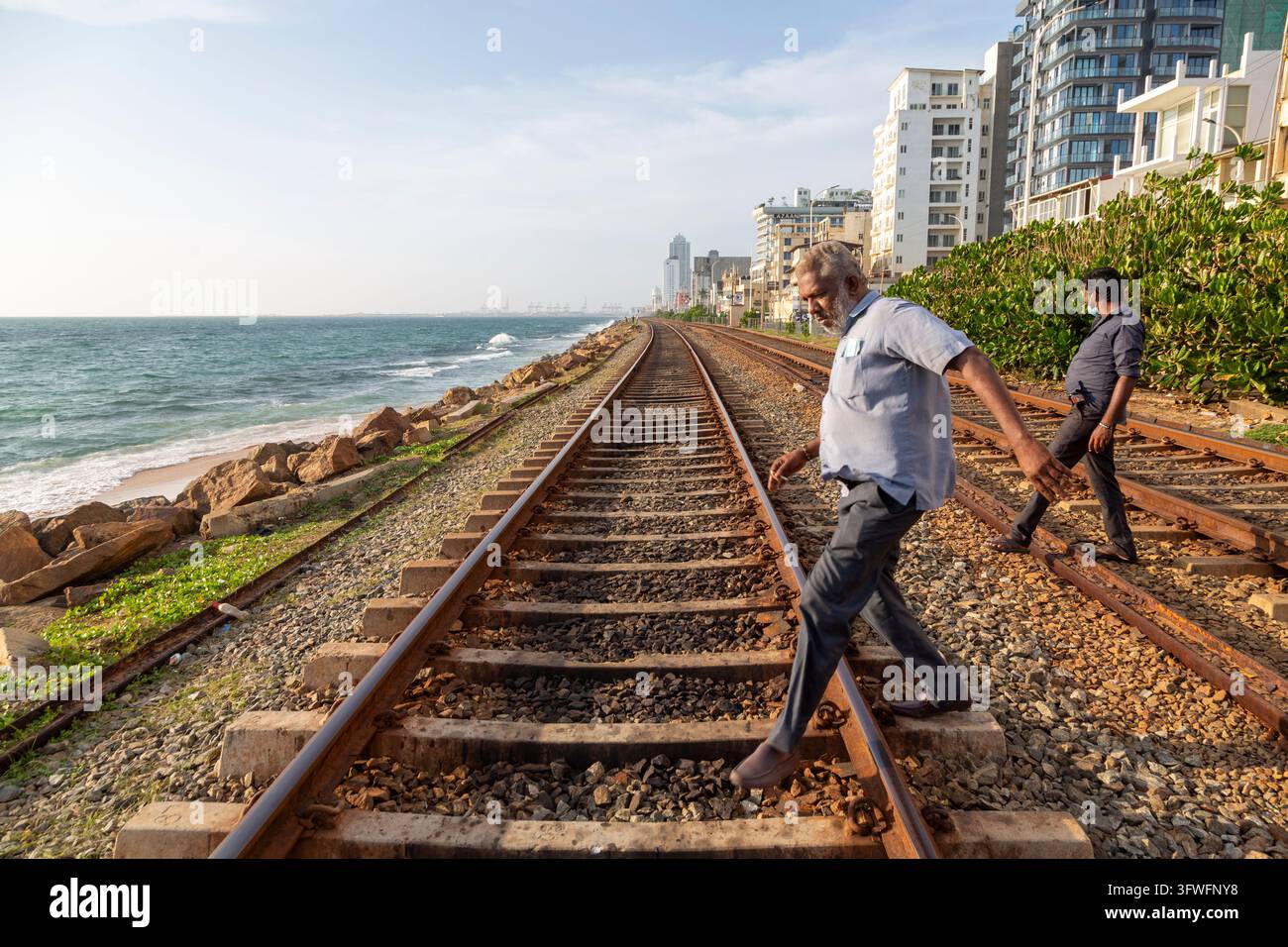 Due uomini attraversano i binari ferroviari accanto al mare e agli edifici residenziali, Provincia occidentale, Colombo, Sri Lanka Foto Stock