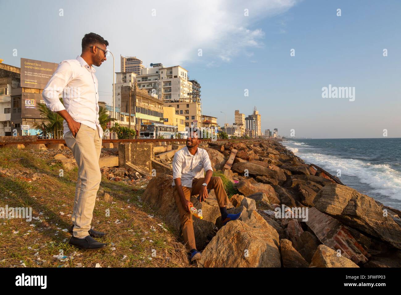 Due giovani uomini chiacchierano sui rocce vicino al mare vicino agli edifici della città, Provincia Occidentale, Colombo, Sri Lanka Foto Stock
