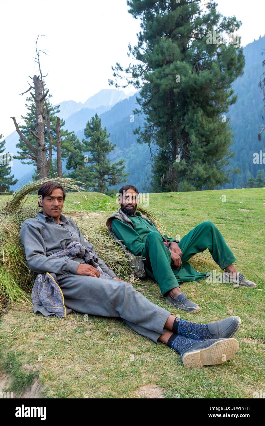 Due uomini nomadi Gujjar che riposano sull'erba contro fasci di erba tagliata, Jammu e Kashmir, valle di Aru, India Foto Stock