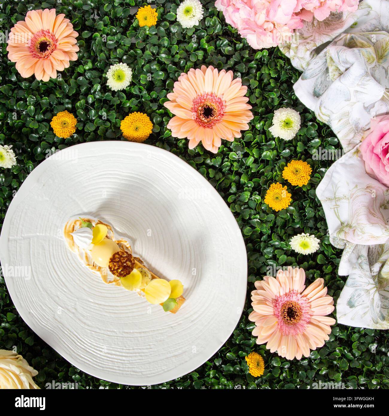 Dessert impiattato con sfere gialle e noci su fogliame verde, circondato da fiori rosa e gialli. Foto Stock